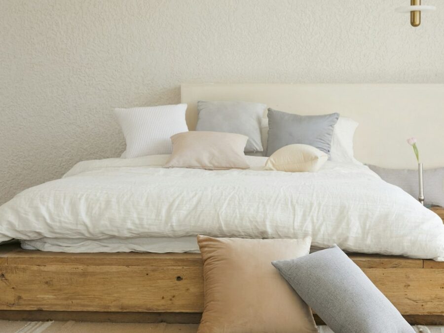 Seng med sengeramme i lyst træ, hvidt sengetøj og puder i lyse farver